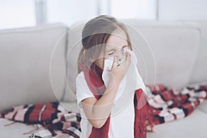 Little sick girl sits on a white couch wrapped in a red scarf. She blows her nose into a napkin