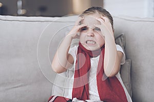 Little sick girl sits on a white couch wrapped in a red scarf. She has a headache and a very high fever