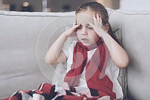 Little sick girl sits on a white couch wrapped in a red scarf. She has a headache and a very high fever