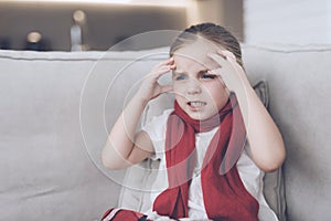 Little sick girl sits on a white couch wrapped in a red scarf. She has a headache and a very high fever