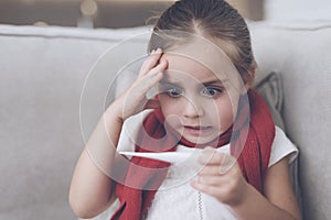 Little sick girl sits on a white couch wrapped in a red scarf. He looks at the thermometer, it has a high temperature