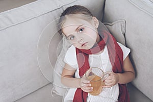 Little sick girl sits on a white couch wrapped in a red scarf. She is sitting with a cup of medicinal tea