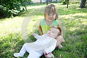 Little sisters in the backyard. Smiling kids sitting on grass in summer. Children in family: toddler and baby portrait