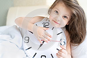 Little sleeping girl with big, huge clock in hands. Early morning wake up before kindergaten,school. White pillow, blanket in bed