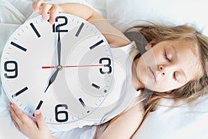 Little sleeping girl with big, huge clock in hands. Early morning wake up before kindergaten,school. White pillow, blanket in bed