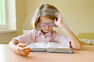 Little smart girl with glasses reading a large book sitting at her desk. School, education, knowledge and children
