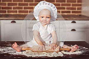 Little smiling baby girl baker in white cook hat and apron kneads a dough