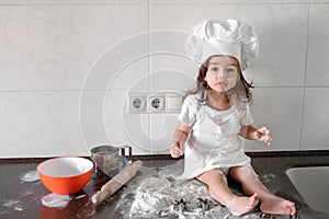 Little smiling baby girl baker in white cook hat and apron kneads a dough on tle kitchen