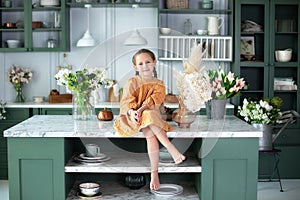 Little Smiling beautiful girl in yellow dress sit at the kitchen island at home. Happy child girl chef on cozy cuisine.