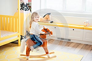 Little smiling blonde 3 years old girl sitting on rocking horse in yellow bedroom at home in day time. Time to play during self