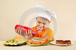 Little smiling boy in chefs hat puts sauce on hamburger