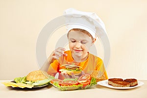 Little smiling boy in chefs hat puts tomato on hamburger