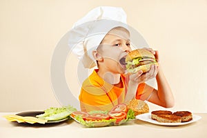 Little smiling boy in chefs hat is tasting cooked hamburger