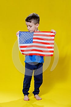 Little smiling boy holding an American flag on a yellow background. Patriotism, independence day, flag day concept