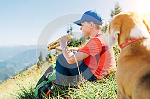 Little smiling boy weared baseball cap enjoying a huge baguette sandwich and his beagle dog friend watching it during a mountain