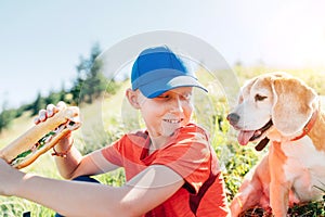 Little smiling boy weared baseball cap with a huge baguette sandwich with his beagle dog friend during a mountain hiking resting