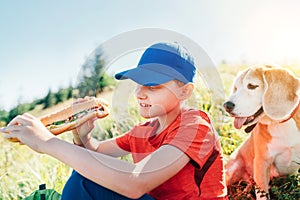 Little smiling boy weared baseball cap with a huge baguette sandwich with his beagle dog friend during a mountain hiking resting