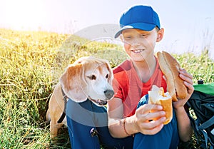 Little smiling boy weared baseball cap sharing a huge baguette sandwich with his beagle dog friend during a mountain hiking