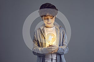 Little smiling curly boy stands on a gray background and looks at a bright light bulb in his hands.