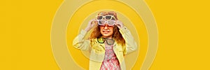 Little smiling cute girl, child with curly hair posing with many sunglasses over yellow studio background. Concept of