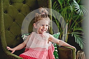 Little smiling girl in a beautiful dress on a green chair.