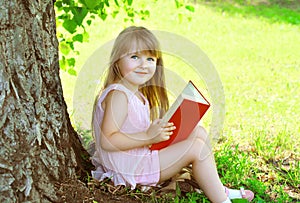 Little smiling girl child reading a book on the grass near tree