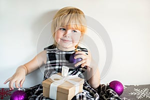 Little smiling girl with Christmas gift box and decorative balls in hands sitting on the bed in dress against white wall