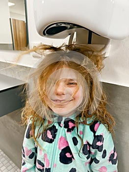 Little smiling girl dries hairs with hairdryer after vistining swimming pool. Happy child with long blond hairs.