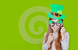 Little smiling girl in green hat of leprechaun and shamrock clover glasses for irish St. Patrick& x27;s Day on green