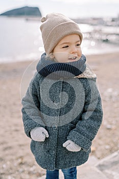 Little smiling girl in mittens stands on the beach looking to the side