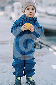 Little smiling girl in overalls with ripe persimmon stands on the pier
