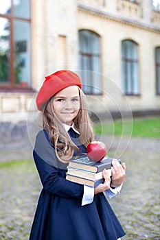 Little smiling girl in a red beret holds a stack of books with an apple near school building. Cute schoolgirl of primary school on