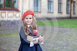 Little smiling girl in a red beret holds a stack of books with an apple near school building. Cute schoolgirl of primary school on