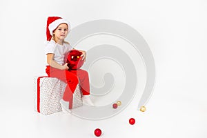 Little smiling girl in santa helper hat sitting on a giftbox with red ribbon and holding red christmas ball, white isolated