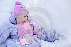 Little smiling girl in scarf and hat lies on snow at winter