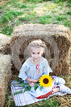 Little smiling girl sits on a roll of haystack in garden and holds a sunflower. A child sits on a straw and enjoys nature in count