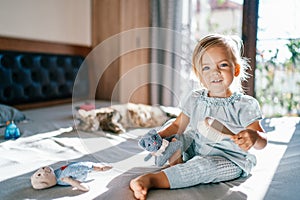 Little smiling girl sitting on the bed with a toy cat and a comb in her hands