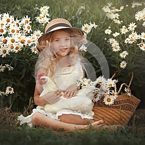 A little smiling girl in a straw hat with a chicken sits under a chamomile bush. Country girl with a basket of flowers