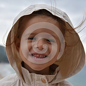 A little smiling girl is wearing a raincoat jacket with hood against the stormy sky. Childhood concepts. Environmental concepts.