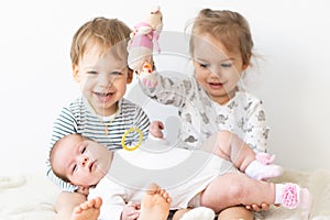 Little smiling kids playing together sitting on the bed. Brother and sister show a newborn a toy. Toddler kids meeting