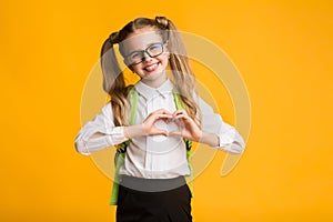 Little smiling schoolgirl showing heart with hands on yellow background