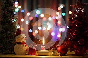 Little snow man with lit red candle light Christmas scenic on wooden table and colorful bokeh background