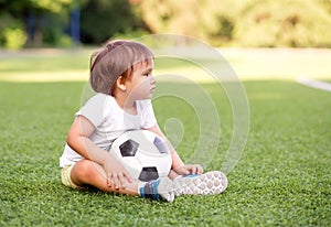 Little soccer fan sitting at football field with ball, looking at stadium. Side view. Toddler boy watching game with serious face
