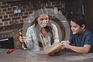 Little son and his smiling mother crafting a wooden birdhouse in kitchen