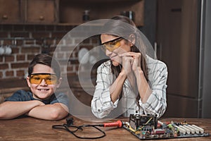 Little son and his young mother in protective glasses sitting at table with motherboard