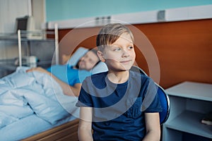 Little son sitting on chair near his sick father sleeping on hospital bed at ward