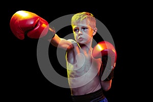 Little sportive boy, beginner boxer in red boxer gloves and shorts training isolated on dark background. Concept of