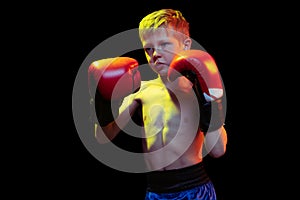 Little sportive boy, beginner boxer in red boxer gloves and shorts training isolated on dark background. Concept of