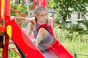 Little sportive girl playing on playground, sitting on the slide