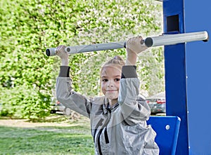 A little sporty girl in a sports uniform is engaged in the exercise machines on the playground for fitness on the street